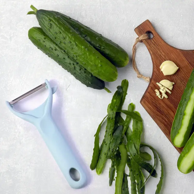 Peeler shown peeling cucumbers next to a wooden board with chopped garlic